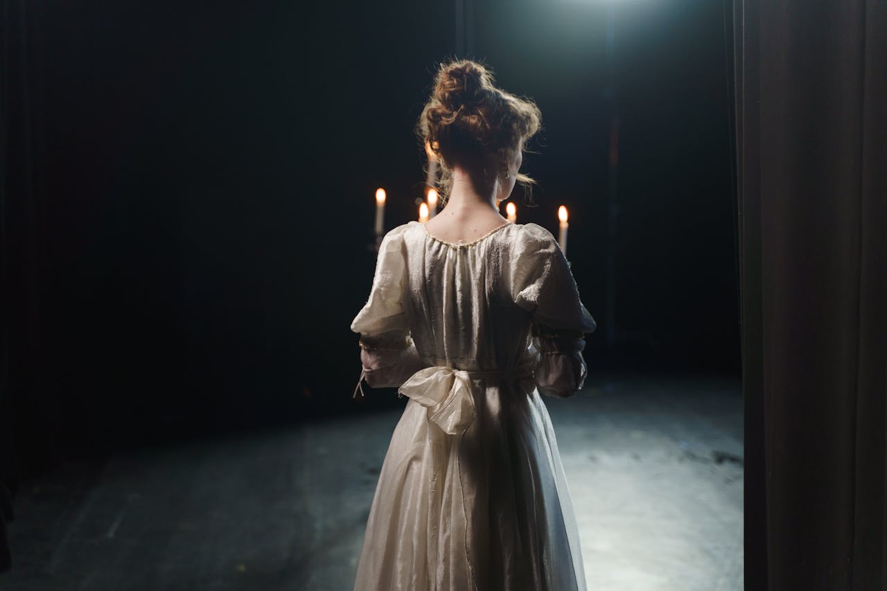 home-hero-1 An actress in a white dress stands backstage in dim lighting, enhancing a classic theatrical atmosphere.