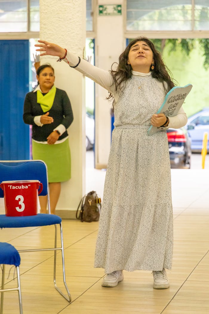 about-us-01 Woman passionately performing indoors with audience in background. Colorful setting with chairs.